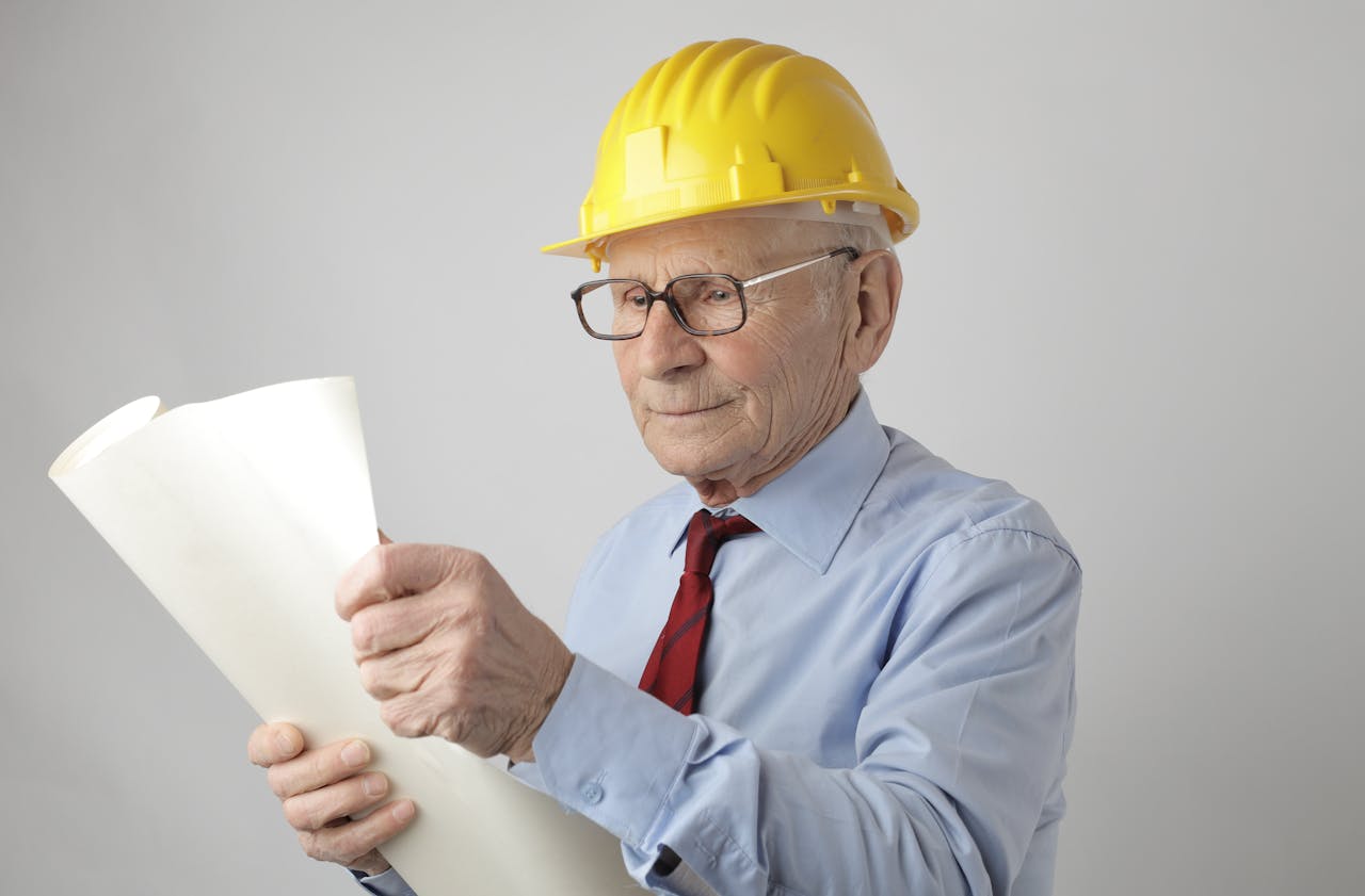 Elderly male engineer in hard hat reviewing blueprints in studio setting.